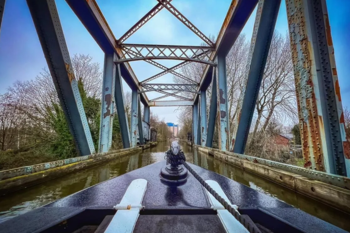 Boat approaching a rusted metal bridge over a canal on a cloudy day.