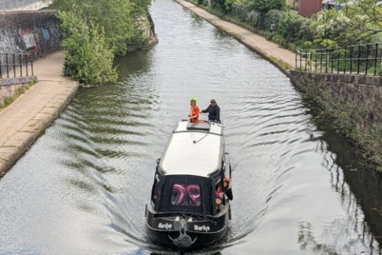 Narrowboat on a canal with two people, surrounded by greenery and distant city buildings.