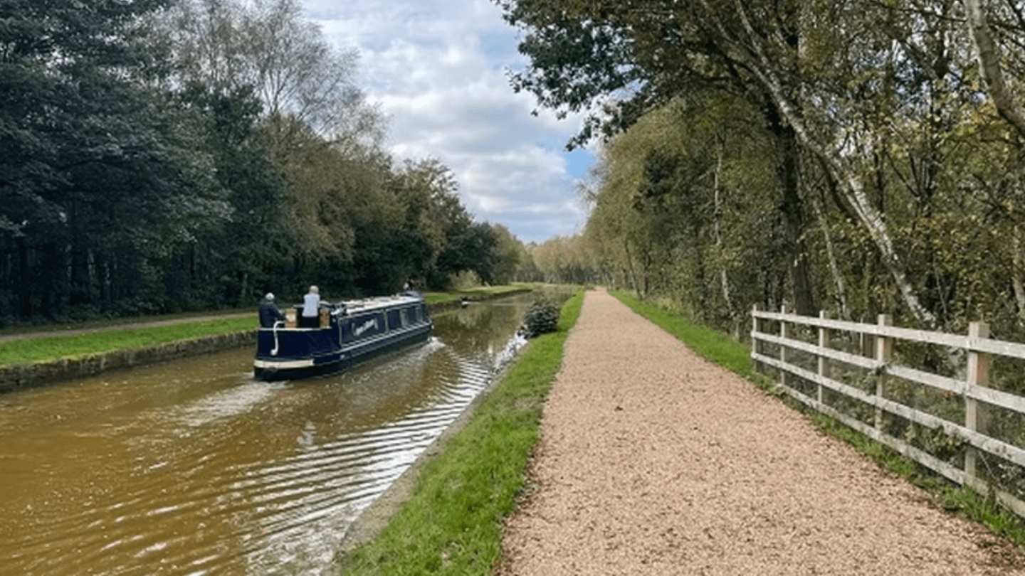 Narrowboat on canal beside gravel path, lined with trees.