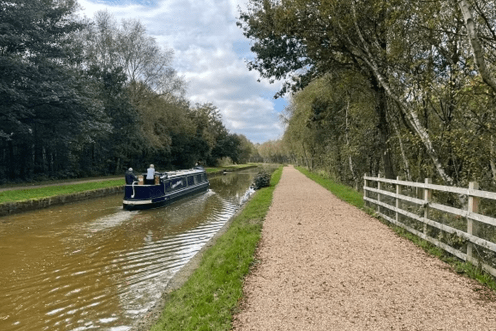 Narrowboat on canal beside gravel path, lined with trees.