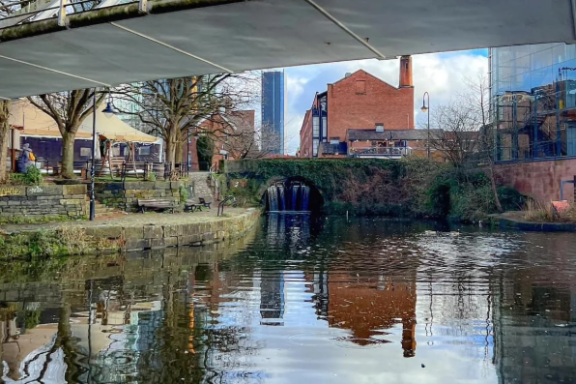 Urban canal scene with bridge, buildings, and trees reflecting in water.