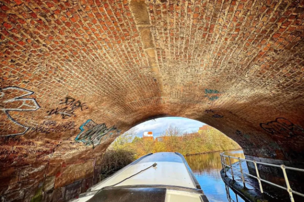 Boat passing under an arched brick bridge with graffiti, leading to a sunny canal scene.