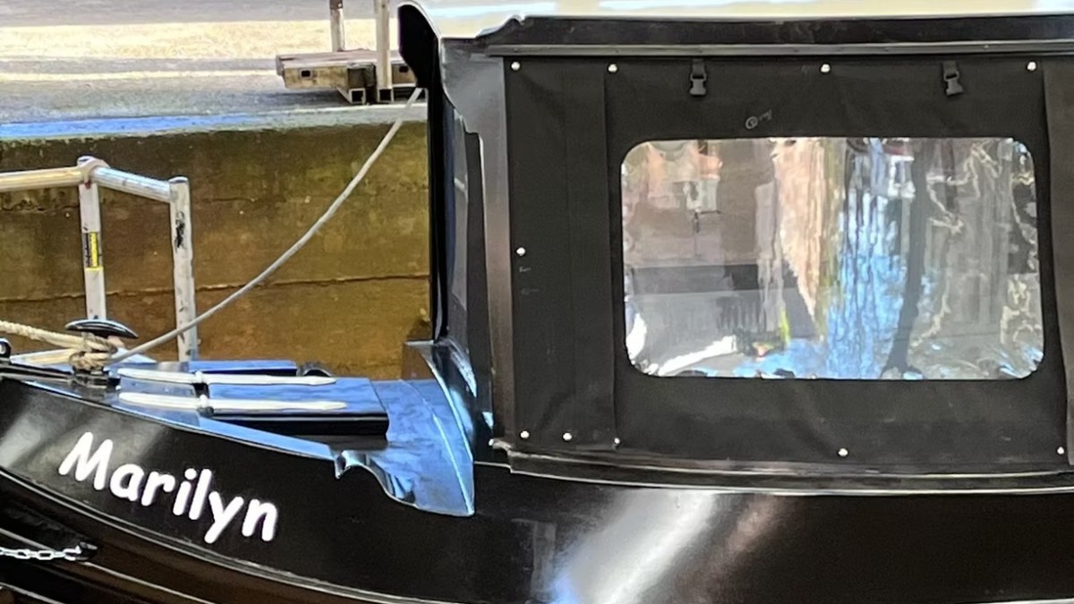 Black boat named 'Marilyn' docked with covered cabin and clear window.