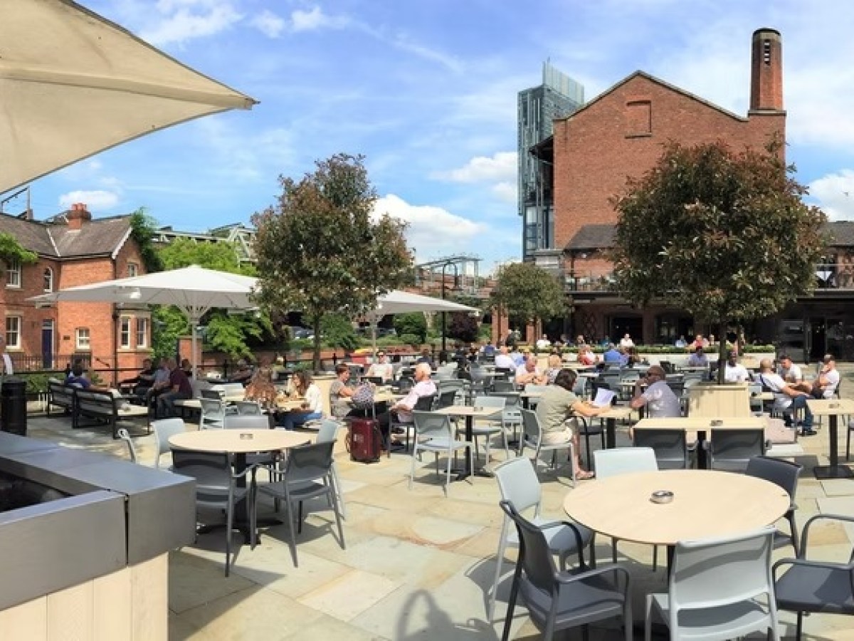 Outdoor cafe with tables, umbrellas, and people, next to a brick building.