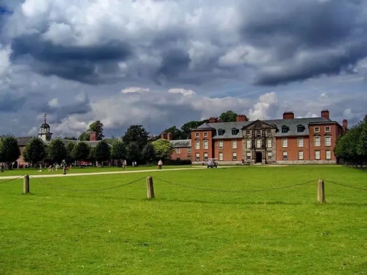 Large brick mansion with chimneys under cloudy sky, surrounded by green lawn and trees.
