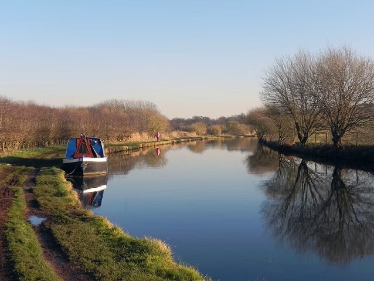 Narrowboat on a calm canal with bare trees reflecting in the water.